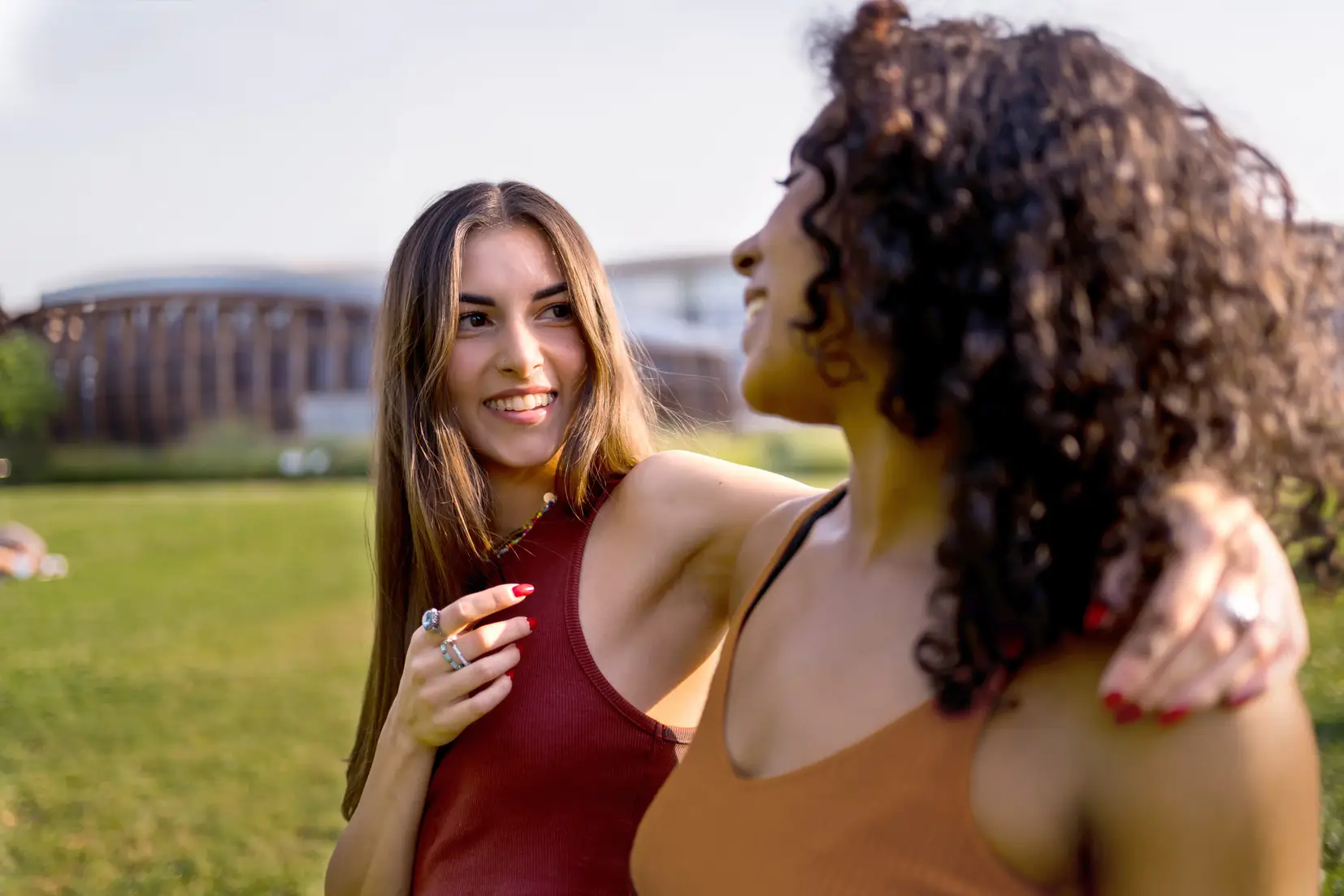 Two women smiling and talking while walking together in a sunny outdoor park setting.